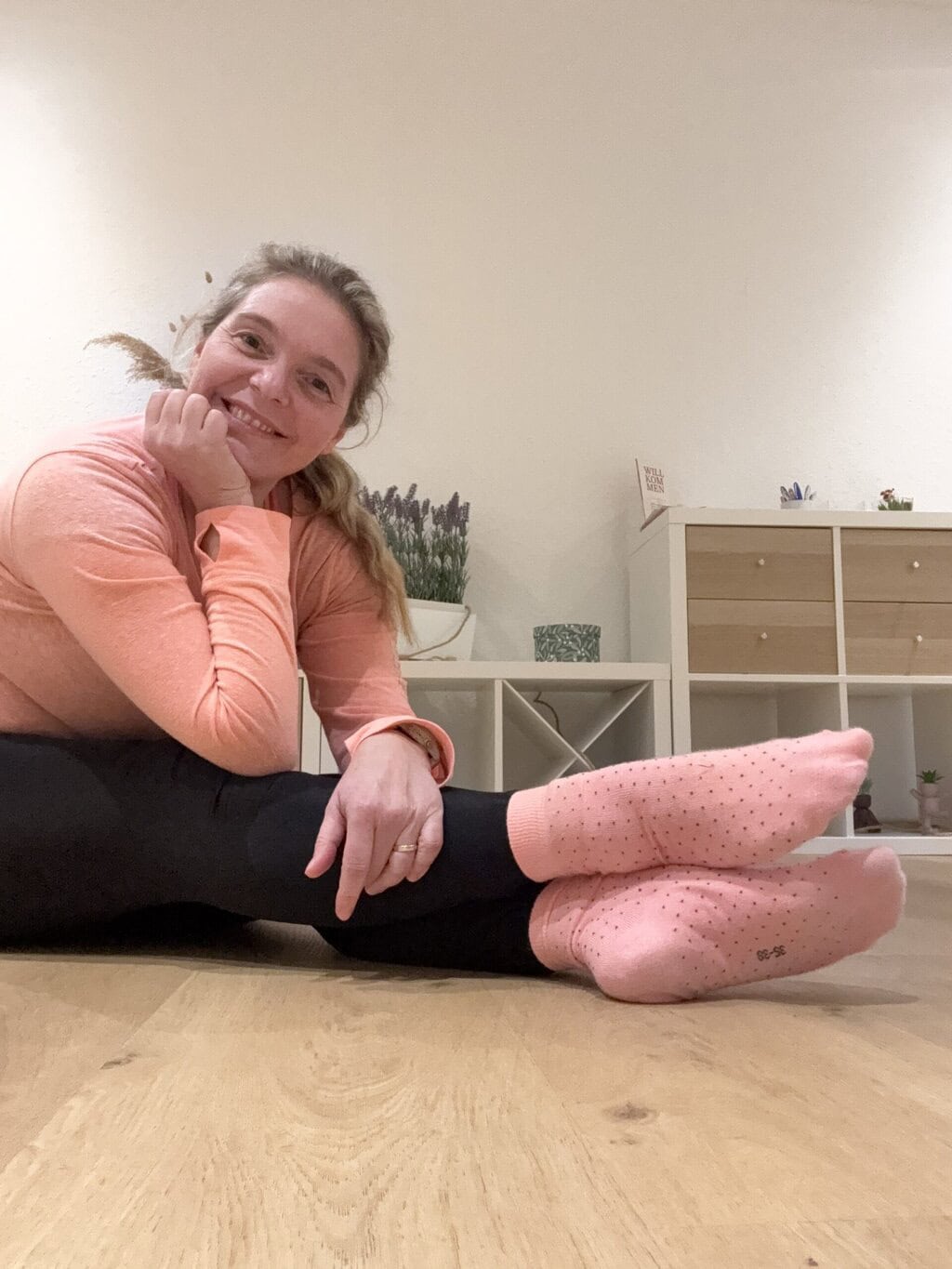 Pilatesanna mit ausgestreckten übereinander geschlagenen Beinen auf dem Boden und lächelt
